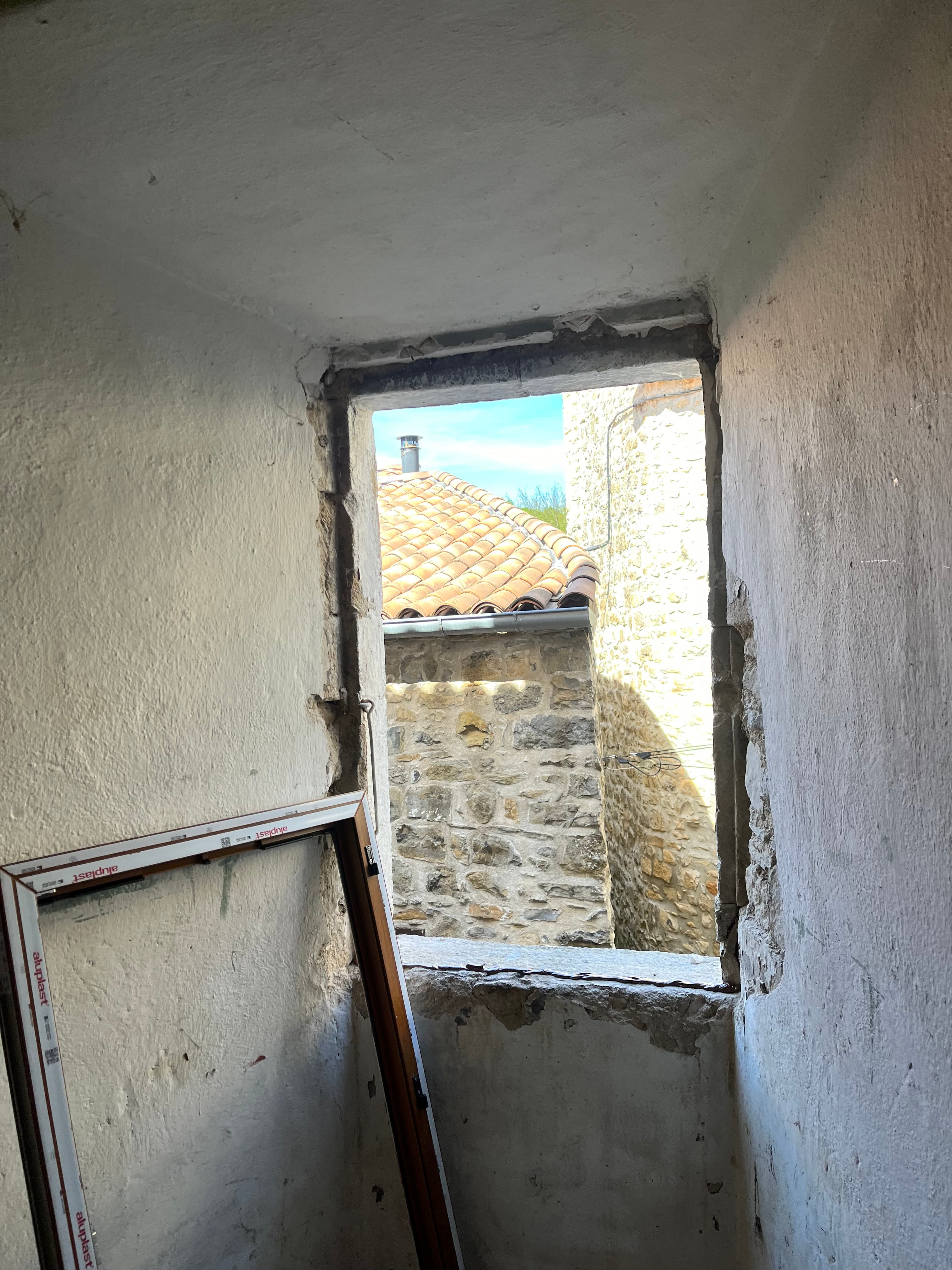 Ouverture de fenêtre brute révélant un toit en tuiles et des murs en pierre ancienne.