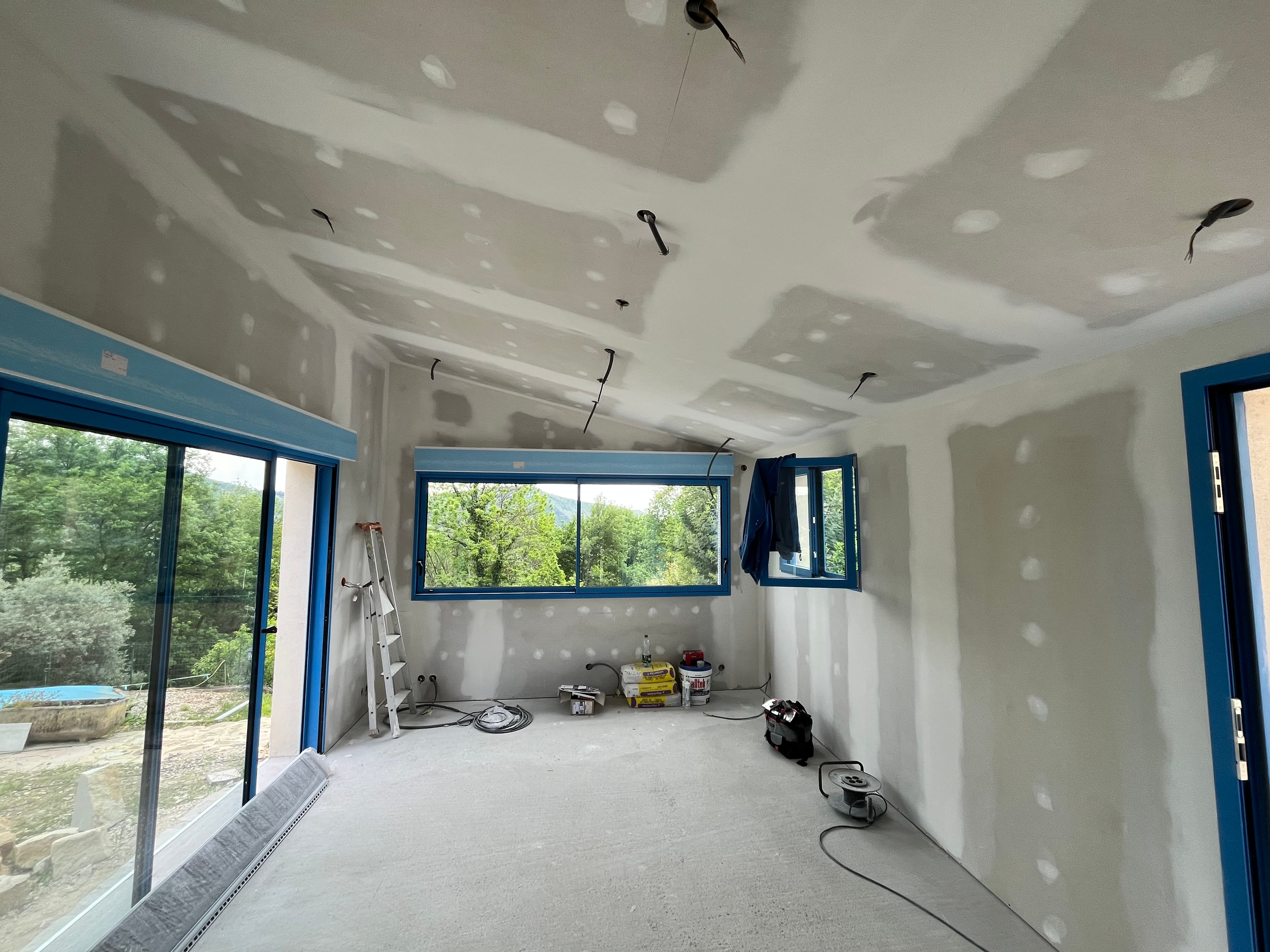 Chantier intérieur avec murs en plaques de plâtre, fenêtres bleues et vue sur la nature.