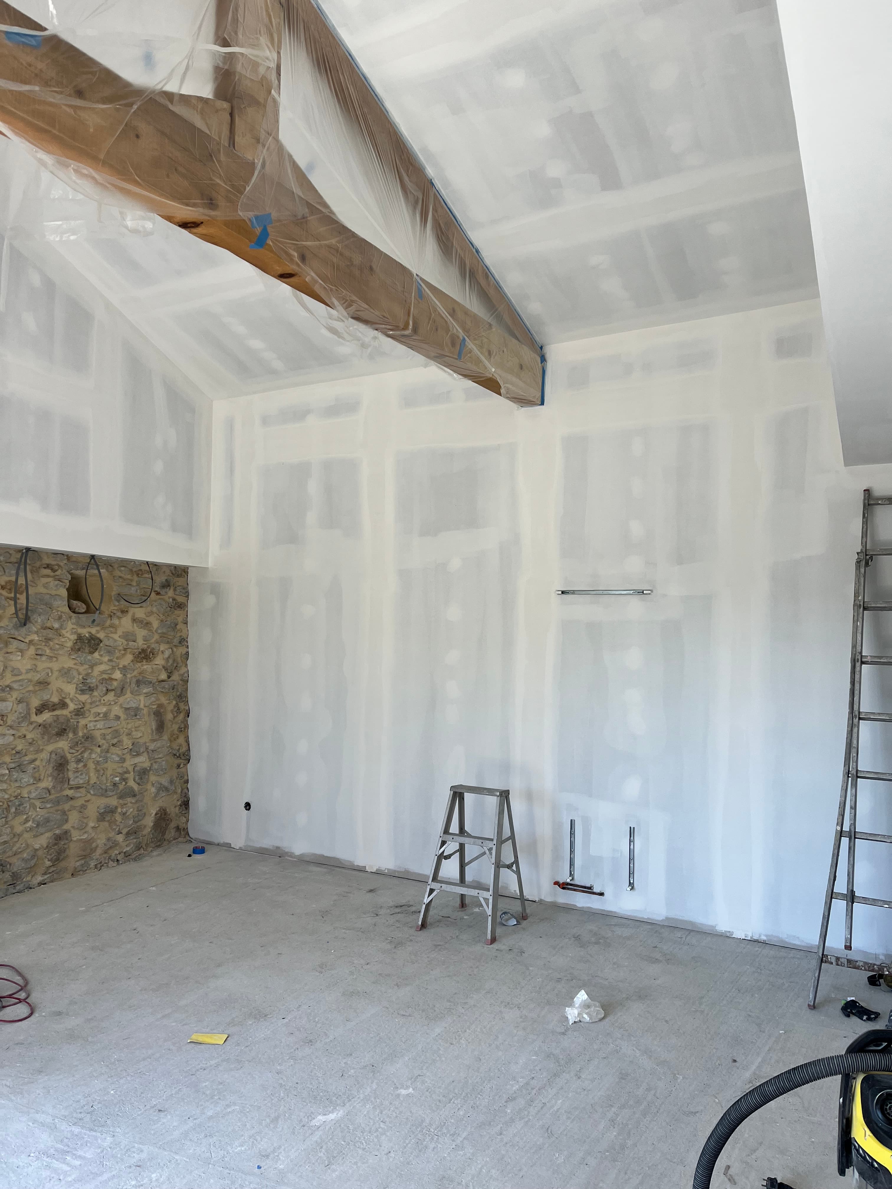 Intérieur en chantier avec plaques de plâtre jointoyées, mur en pierre apparente et échelles.