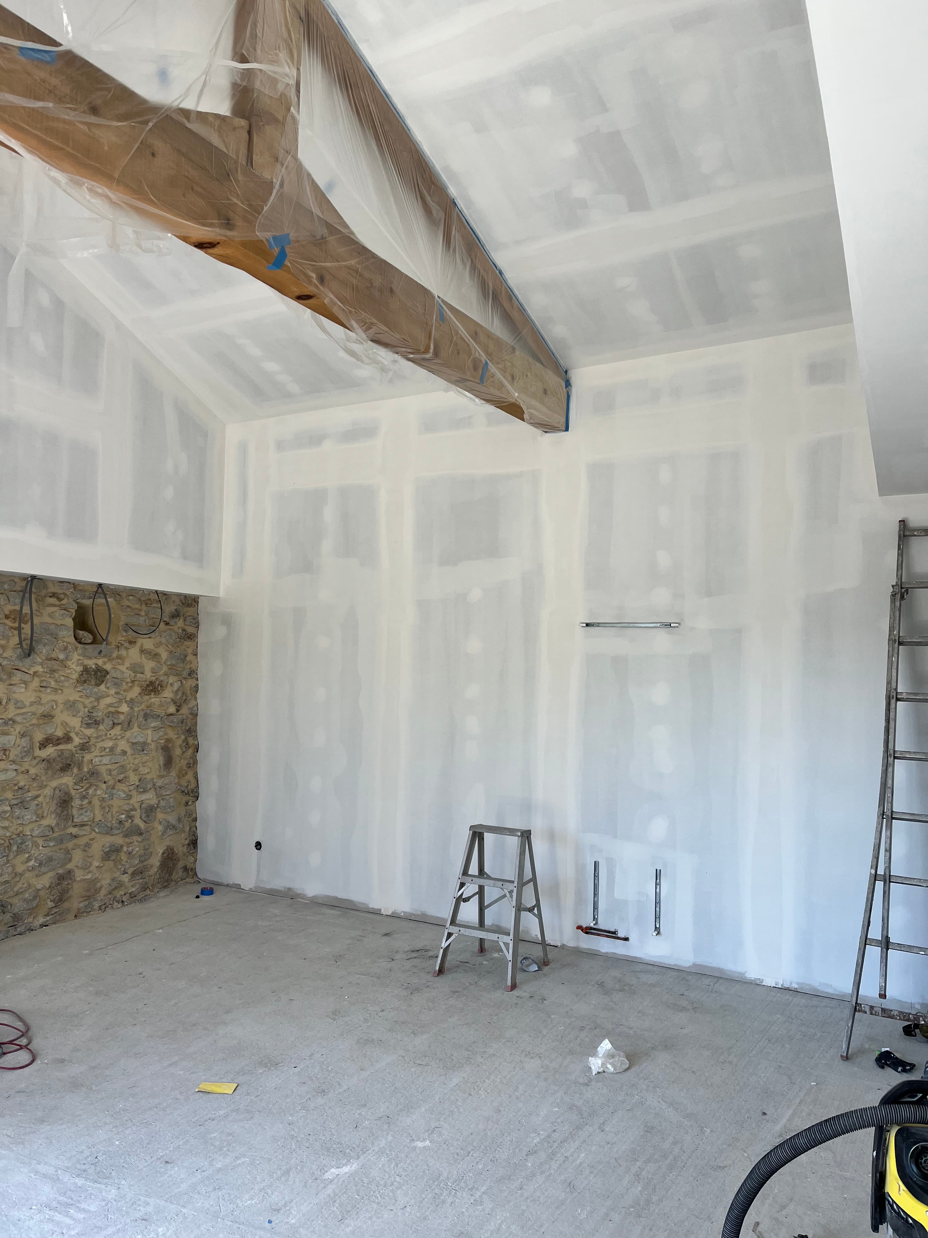 Intérieur en chantier avec plaques de plâtre, poutre en bois protégée et mur en pierre.